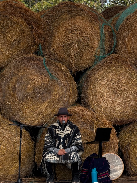 A musician in a poncho and hat in front of a wall of hay bales